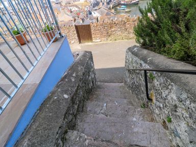 Stone steps leading down to a paved area with a stone wall and wooden gate overlooking rooftops and water at Fishnets in Brixham