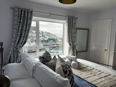 A bedroom with a bed pillows folded towels and patterned curtains by a window overlooking a harbor at Fishnets in Brixham