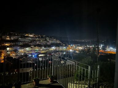 A nighttime view of a harbor with boats and illuminated buildings seen from a balcony with metal railings and potted plants at Fishnets in Brixham