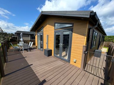 A wooden deck with glass table and chairs outside a modern lodge with double glass doors at Lodge 18 - The Pines in Runswick Bay