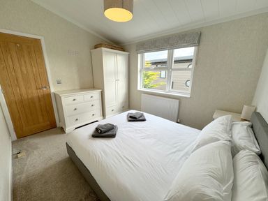 A bedroom with a bed with white linens and towels on top a white wardrobe and chest of drawers a window with a patterned blind and a wooden door at Lodge 18 - The Pines in Runswick Bay
