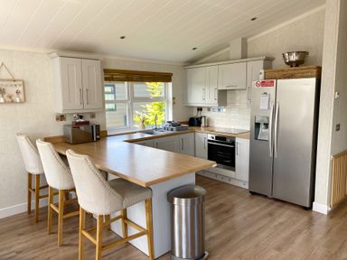 A kitchen with a wooden countertop, three upholstered bar stools, a stainless steel refrigerator, microwave, oven, sink, and cabinets at Lodge 18 - The Pines in Runswick Bay