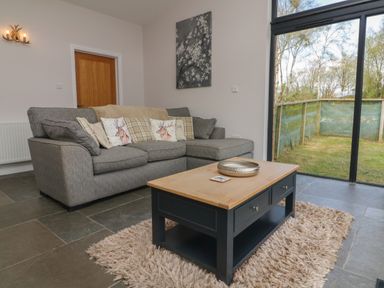 A living room with a grey sofa and patterned cushions a wooden coffee table on a rug and a sliding glass door showing a fenced yard at Silver Birch in Dolton