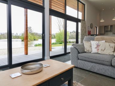 A living room with a grey sofa and deer pillows next to large windows overlooking a lake at Silver Birch in Dolton