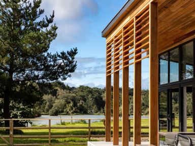 A wooden structure with vertical slats next to glass windows overlooking a lake with trees and grass at Silver Birch in Dolton