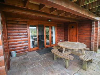 A covered patio area with a round wooden picnic table and benches in front of a wooden cabin with glass doors at Aspen Dolton