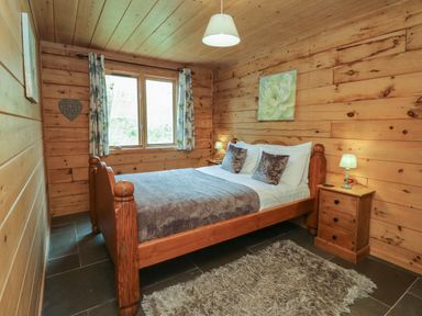 A bedroom with wooden walls and ceiling a wooden bed with pillows and blanket two bedside tables with lamps a window with curtains and a rug on the floor at Aspen in Dolton