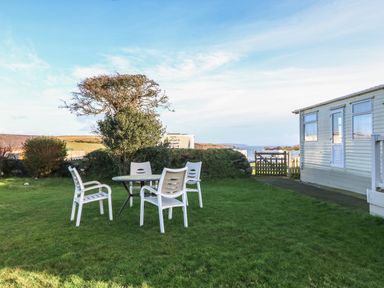 A garden with white plastic chairs and a table next to a caravan and a gate with a view of the coast at Tir Glyn Caravan in Uwch Mynydd near Aberdaron