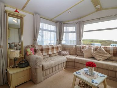 A living room with a beige sectional sofa a small side table with a lamp and radio and a coffee table with a flower vase at Tir Glyn Caravan in Uwch Mynydd near Aberdaron
