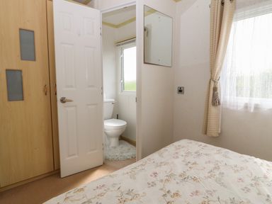 A bedroom with a floral bedspread a window with curtains a mirror on the wall a wardrobe and a partially open door showing a toilet with a window at Tir Glyn Caravan in Uwch Mynydd near Aberdaron