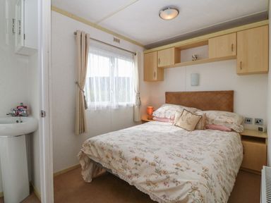 A bedroom with a double bed floral bedding wooden cabinets above and beside the bed a window with curtains and a small sink area with toiletries at Tir Glyn Caravan in Uwch Mynydd near Aberdaron