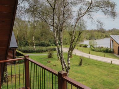 A balcony overlooking trees a grassy area a gravel path and a lake with a wooden building nearby at Cedar in Dolton