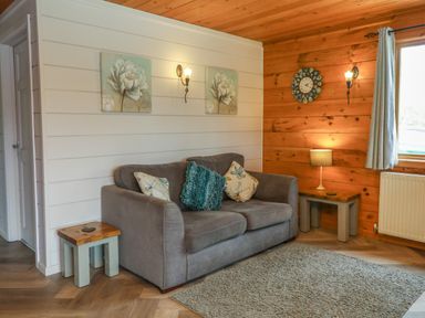 A living room with a grey sofa cushions two wooden side tables a lamp wall clock and two floral paintings at Cedar in Dolton