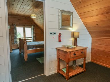 A wooden bedside table with a lamp and a mirror above it next to a doorway leading to a bedroom with a wooden bed at Cedar in Dolton