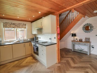 A kitchen with white cabinets wooden ceiling and floor and a small table with a lamp near a wooden staircase at Holly in Dolton