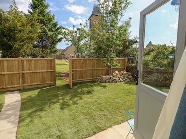A garden with grass wooden fence a small tree and a stone pile visible from an open door at Tower Hut in Ryde
