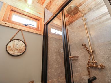 A bathroom shower area with brass shower fixtures a small window and a round mirror hanging on the wall at Tower Hut in Ryde