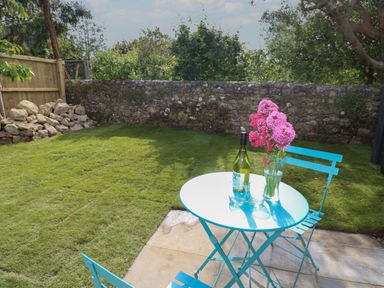 A garden with a blue metal table and chairs on a stone patio a bottle of wine two glasses and a vase with pink flowers on the table at Tower Hut in Ryde
