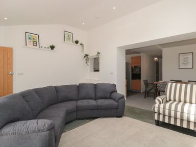 A living room with a curved dark gray sofa a striped armchair and shelves with framed pictures and plants at Primrose Cottage in Brendon Hill near Roadwater
