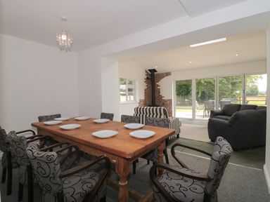 A dining room with a wooden table set with white plates and patterned chairs adjacent to a living area with sofas and a wood stove at Primrose Cottage in Brendon Hill near Roadwater