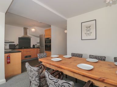 A dining room with a wooden table and gray patterned chairs next to a kitchen with wooden cabinets and stainless steel appliances at Primrose Cottage in Brendon Hill near Roadwater