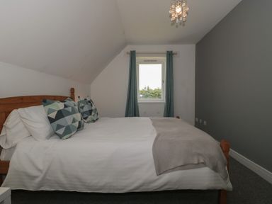 A bedroom with a wooden bed white bedding two patterned pillows a gray blanket a window with curtains and a small ceiling light at Primrose Cottage in Brendon Hill near Roadwater