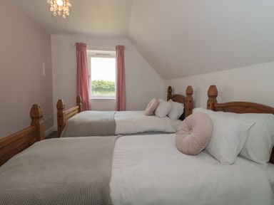 A bedroom with two single beds with wooden frames and pink round cushions near a window with pink curtains at Primrose Cottage in Brendon Hill near Roadwater