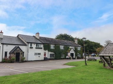 A white building with ivy on the walls surrounded by a parking lot and green grass with picnic tables and lamp posts at Primrose Cottage in Brendon Hill near Roadwater