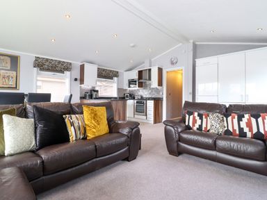 A living room with two brown leather sofas with cushions and an open kitchen area with white cabinets and appliances at Capenwray in Carnforth