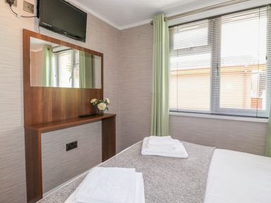 A bedroom with a bed containing folded towels a wall-mounted television a wooden shelf with a mirror and a vase of flowers near a window with blinds and green curtains at Capenwray in Carnforth