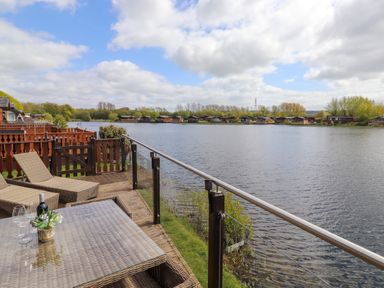 A balcony with rattan furniture overlooking a lake with wooden cabins on the shore at Capenwray in Carnforth