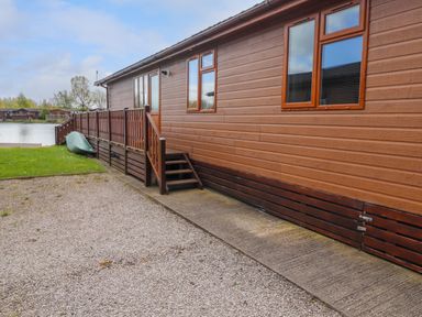 An exterior side view of a wooden cabin with a small staircase and a railing next to a gravel driveway and a grassy area near a body of water at Capenwray in Carnforth