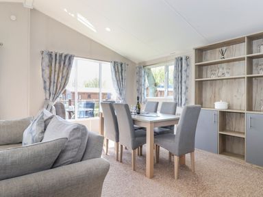 A dining area with a wooden table and six grey upholstered chairs next to windows with patterned curtains and a shelving unit with glassware and a white pot at Coniston in Carnforth