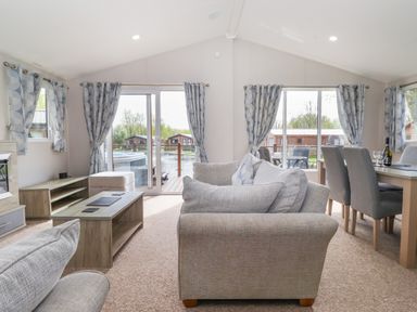 A living room with gray sofas a wooden coffee table and a dining table with chairs near large windows with curtains at Coniston in Carnforth