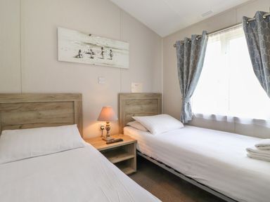 A bedroom with two single beds a wooden nightstand with a lamp and remote between the beds and a window with curtains at Coniston in Carnforth