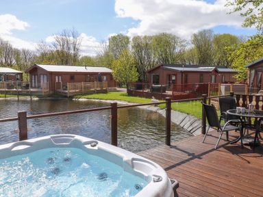 A deck with a hot tub and table with chairs overlooking a pond and wooden cabins surrounded by trees at Coniston in Carnforth