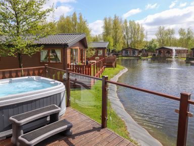 A deck with a hot tub overlooking a pond with multiple wooden cabins and trees at Coniston in Carnforth