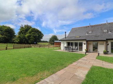 A backyard with a lawn and patio furniture next to a house with a paved pathway at Campion Cottage in Brendon Hill near Roadwater