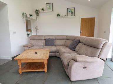 A living room with a beige sectional sofa wooden coffee table and floating shelves with plants and framed pictures at Campion Cottage in Brendon Hill near Roadwater