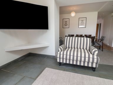 A room with a striped armchair a wall-mounted TV a corner shelf a dining table with chairs and butterfly framed pictures at Campion Cottage in Brendon Hill near Roadwater