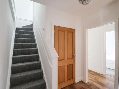 A hallway with carpeted stairs a wooden door and an open doorway showing a bedroom at Campion Cottage in Brendon Hill near Roadwater