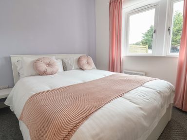 A bedroom with a bed made with white linens and pink cushions near windows with pink curtains at Campion Cottage in Brendon Hill near Roadwater