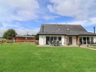 A house with a dark roof and white walls with outdoor chairs and tables on a patio and a green lawn in front at Campion Cottage in Brendon Hill near Roadwater