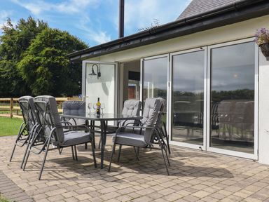 An outdoor patio with a glass table and six cushioned chairs near glass sliding doors at Campion Cottage in Brendon Hill near Roadwater