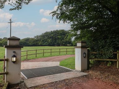 An entrance gate with two white pillars and metal cattle grid leading to a dirt road and grassy field at Campion Cottage in Brendon Hill near Roadwater