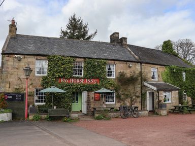 A stone building covered with ivy with a sign reading Bay Horse Inn with outdoor benches and umbrellas