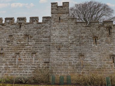 A stone castle wall with battlements and narrow windows with plants and bushes in front at Cragg Lodge in West Woodburn