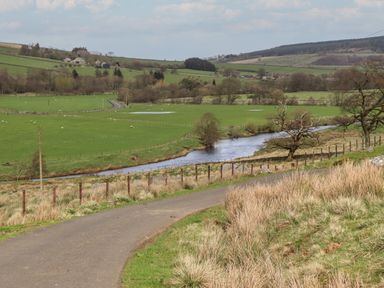 A countryside landscape with a winding river running through green fields and a narrow road in the foreground at Cragg Lodge in West Woodburn