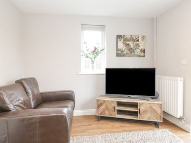 A living room with a brown leather sofa a wooden TV stand with a television a small window with flowers and a painting on the wall at The Brambles in Monmouth