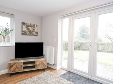 A corner of a living room with a television on a wooden stand a window with flowers and glass doors leading outside at The Brambles in Monmouth